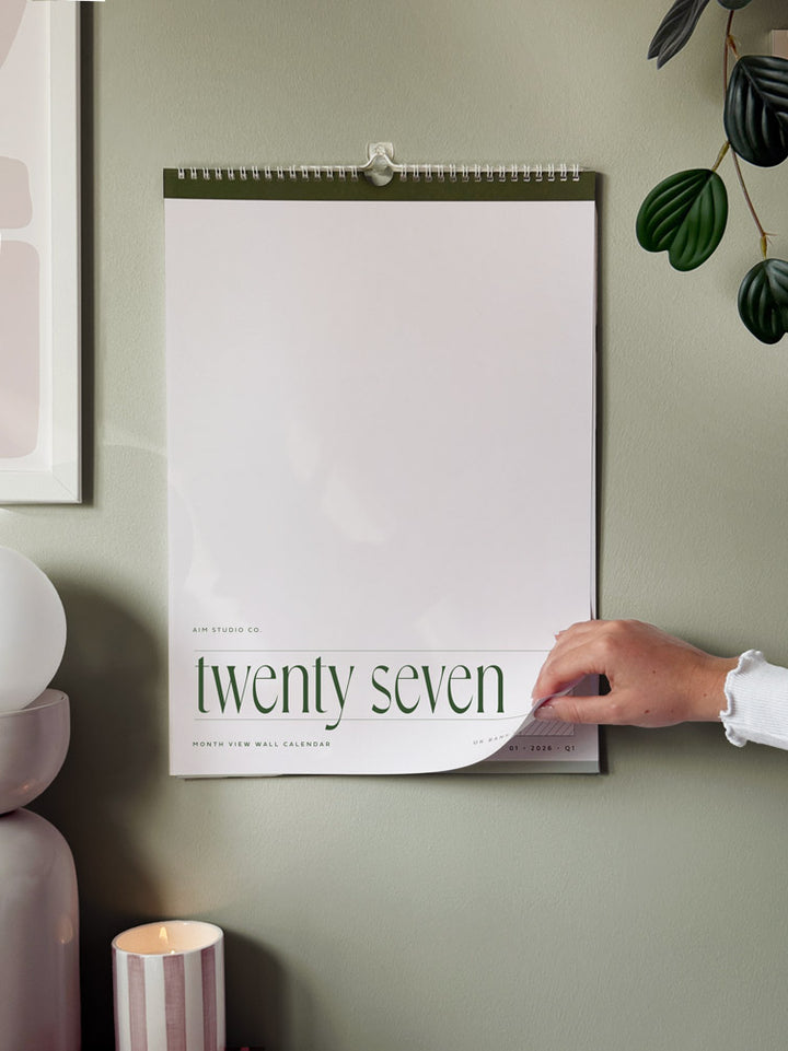 A spiral-bound wall calendar hangs on a muted sage green wall. A hand is peeling back the bottom page, which reads &