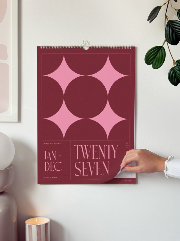 2027 wall calendar with a bold pink and burgundy geometric star design is hanging on a white wall. A hand is peeling back the bottom-right page to reveal the days of the week.