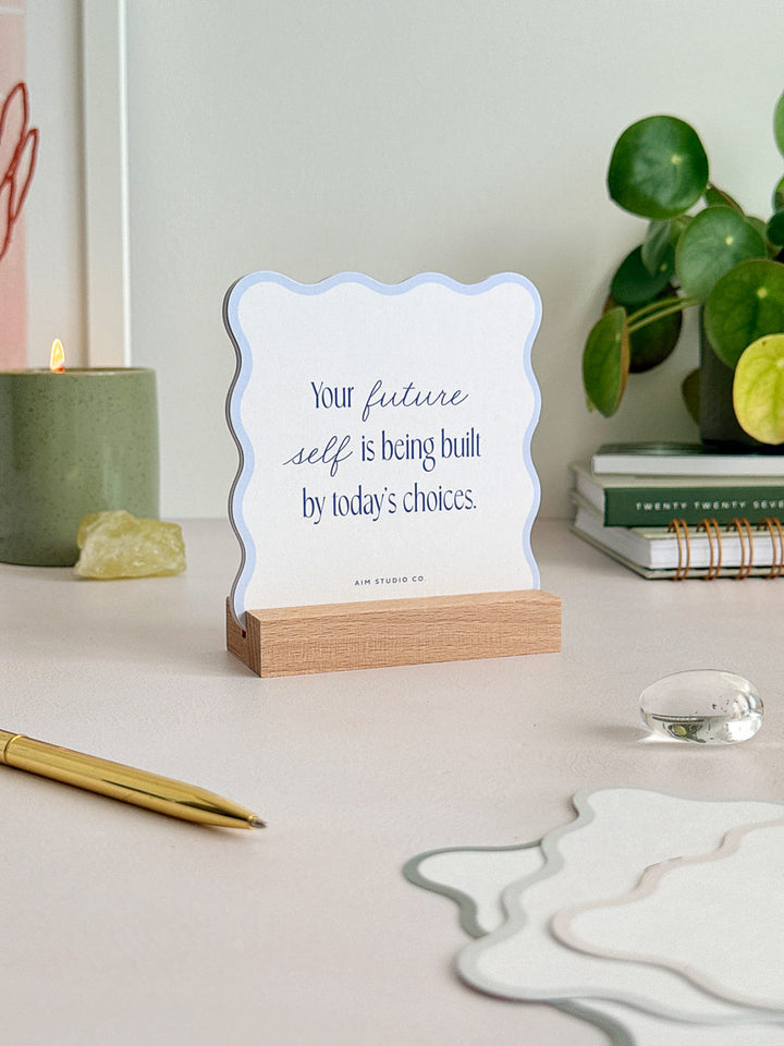 an office desk accessory showing a daily quote card for confidence and personal growth. The card is cut into a wavy shape and has a pale blue scalloped border, held in a wooden holder to display the card upright.