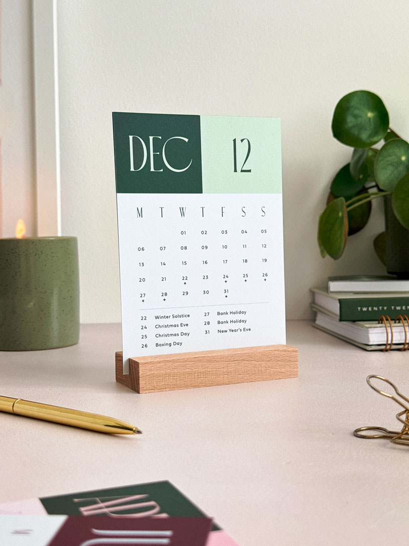 Deep forest green and sage green month to view calendar card held in a wooden holder by AIM Studio Co. The calendar shows decemeber 2027 with UK bank holidays and a minimal monday start grid of dates.
