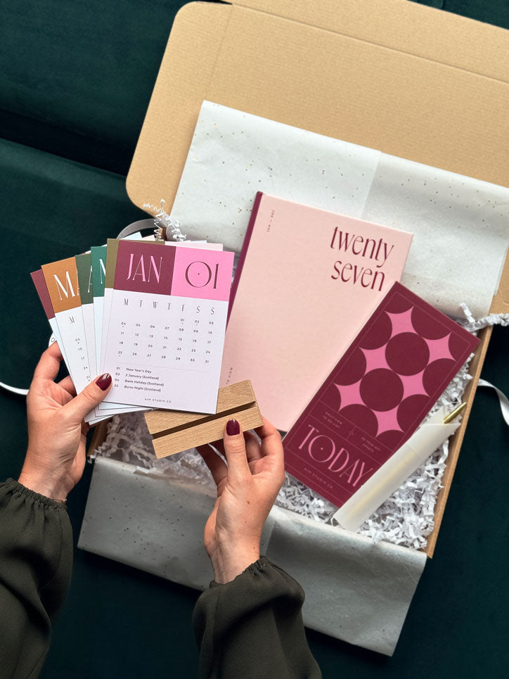 hands holding a 12 month desk calendar card pack with a wooden holder to display them on a desktop. Below, the rest of the stationery gift bundle features a pale pink 2027 diary and a pink and burgundy geometric notepad with pen.