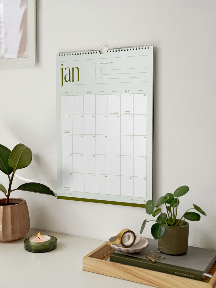 January monthly calendar hanging on a wall showing planner grid with UK bank holidays on a clean minimalist page. Surrounded with green desk accessories and a plant.