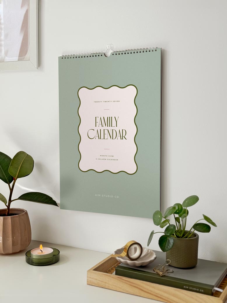 A sage green spiral-bound family calendar hanging on a white wall in a styled home setting, surrounded by potted plants, a candle, and stationery on a desk.