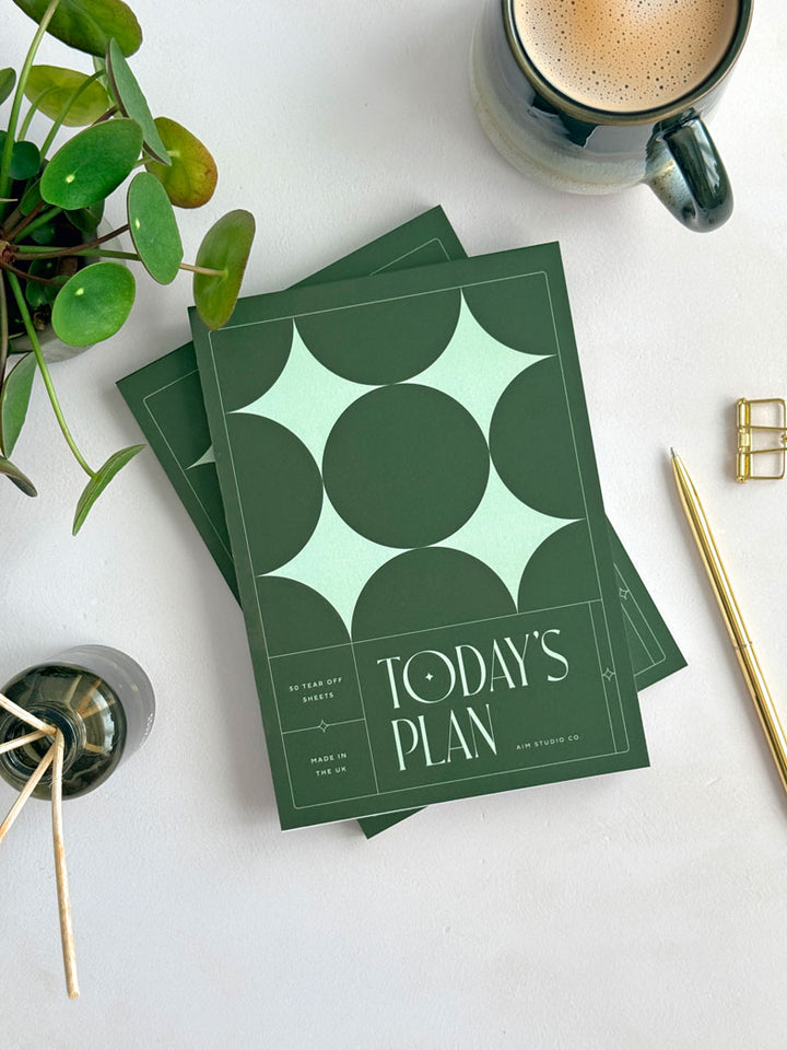 A5 sized daily planner notepad on a modern office desk setting. The cover of the notepad is dark green and features light green stars in a geometric grid layout. Bold serif typography reads &