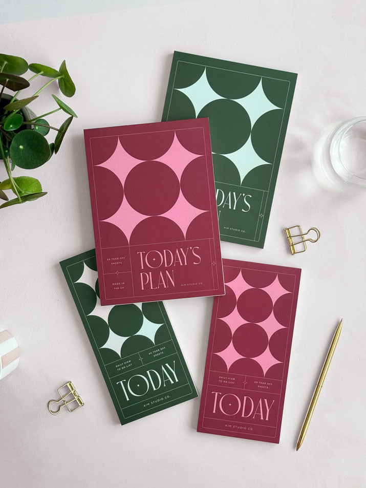 A styled flat-lay showing all 4 daily planner options - two A5 notepads and two slim DL-sized notepads in burgundy and pink or deep green and mint green. The cover designs all feature a modern geometric star pattern with large serif typography reading "today" and "today&