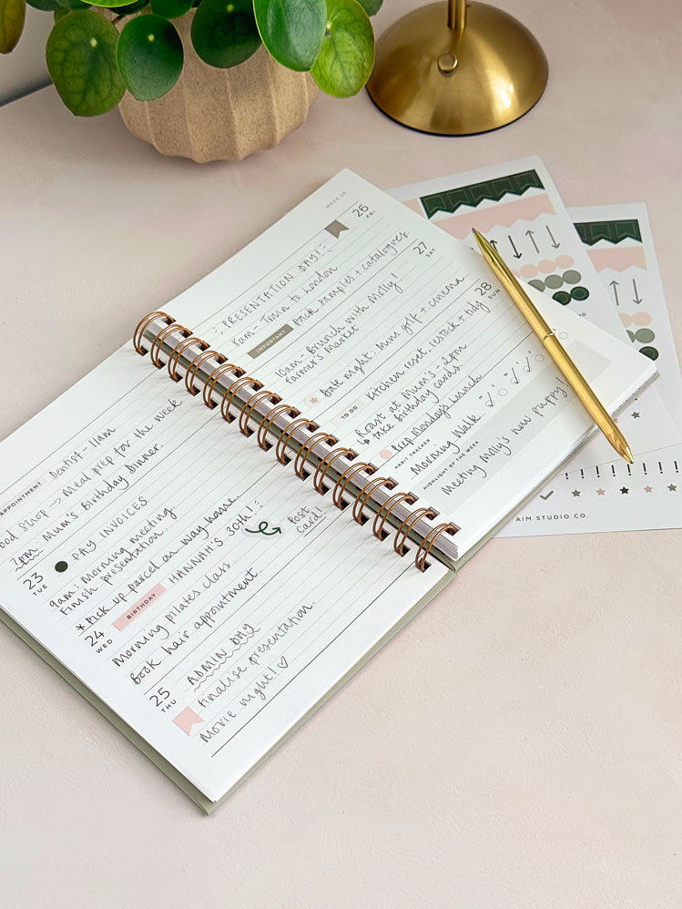 An open spiral-bound week to view diary with handwritten notes lies on an office desk, styled with a gold pen and planner sticker sheets.
