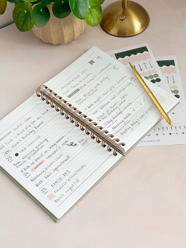 An open spiral-bound week to view diary with handwritten notes lies on an office desk, styled with a gold pen and planner sticker sheets.