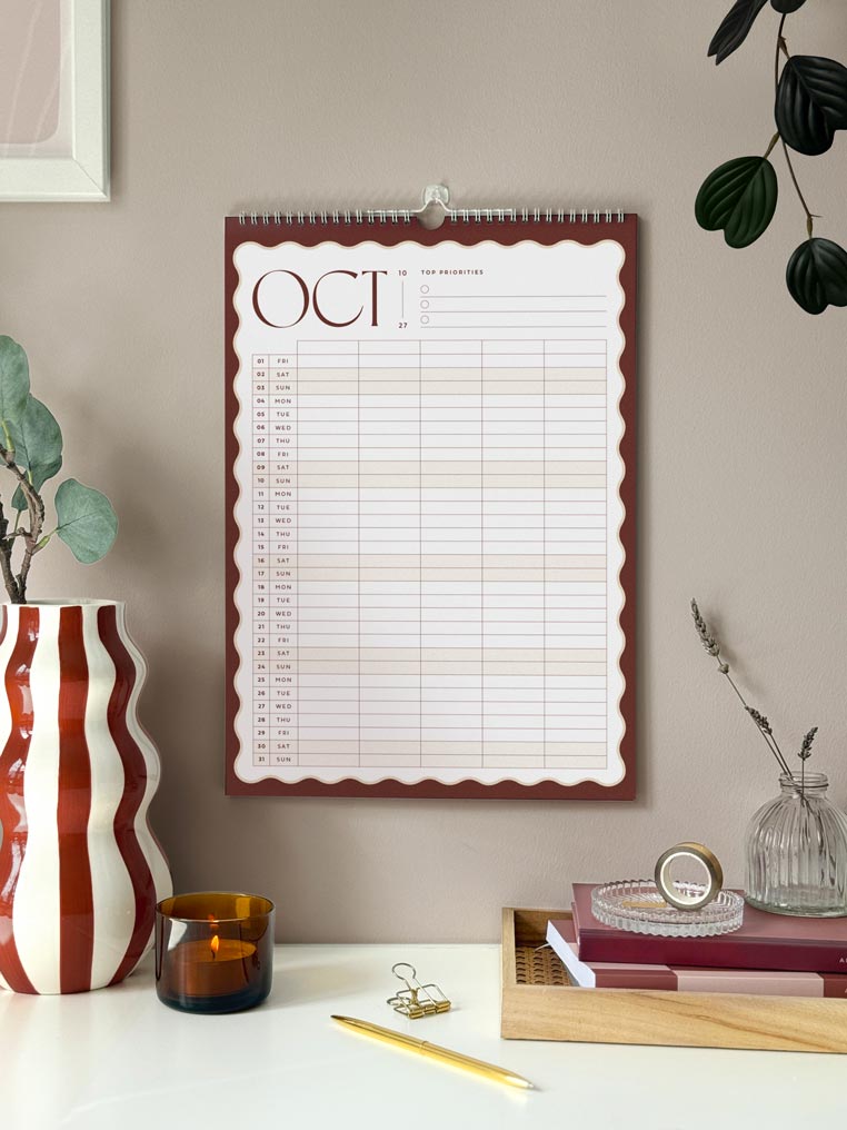 October 2027 monthly page of large family calendar hanging on a brown wall above a desk. the planner shows a month view with scalloped edge in deep brown and a vertical planner grid.