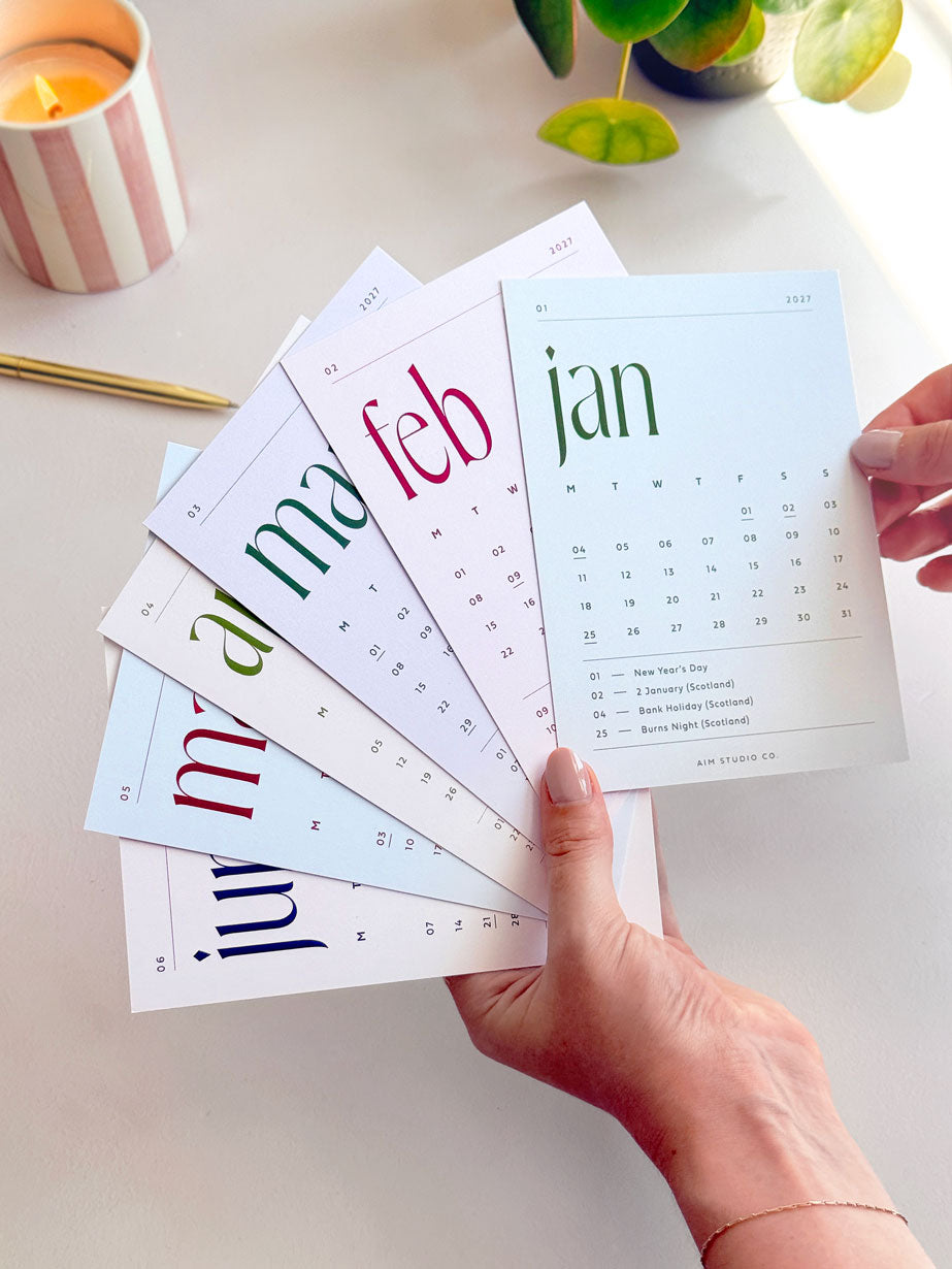a woman holding the full set of 2027 month to view small desk calendar cards in an assorted set of soft neutral tones with bold serif typography.