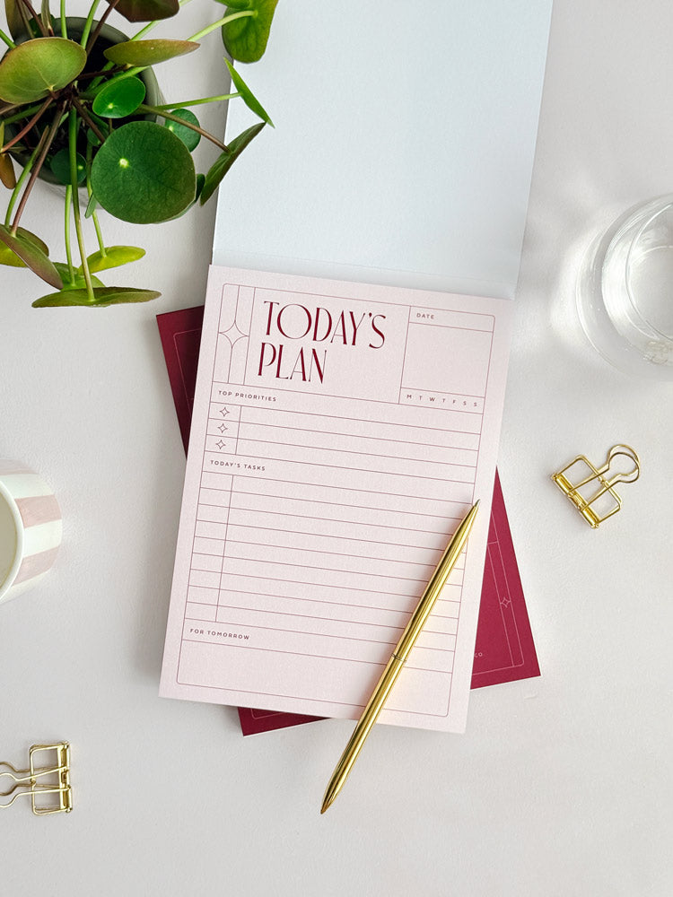 Styled flat-lay of pale pink daily planner notepad with over, surrounded by desk plant, candle and gold stationery accessories.