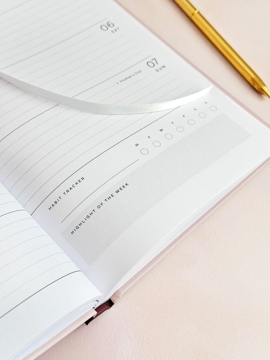 Daily habit tracker section in 2027 weekly planner by AIM Studio Co. The diary is laying open on a weekly planner page with a white ribbon bookmark over the page and gold pen next to it.