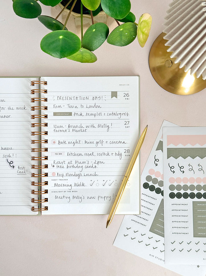 a5 spiral diary with weekly planner spread open showing planner stickers and handwritten plans on the page. Styled on a womans office desk with a gold pen and aesthetic accessories.