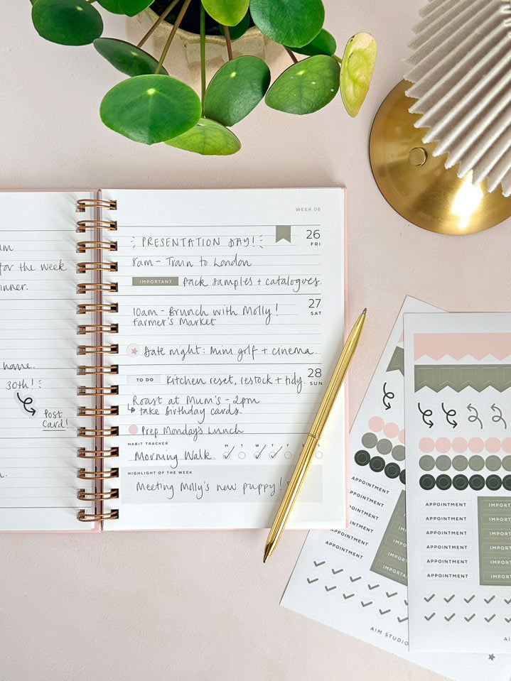 A styled flat-lay of a spiral-bound 2027 diary with handwritten notes, decorated sticker sheets, a gold pen, and green leaves on a woman&