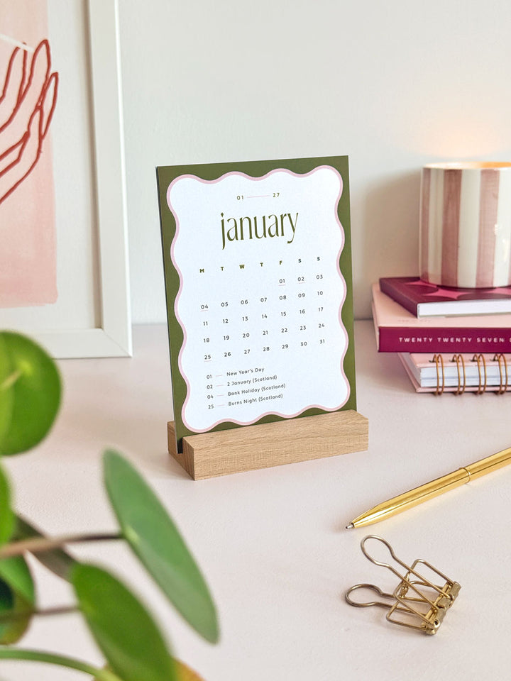 January 2027 desk calendar on an office desk with olive green and contrasting pink scalloped border. The calendar card is displayed in a wooden block on a desktop surface, styled with aesthetic desk accessories for women.