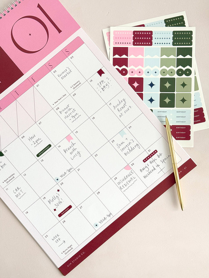 A planner page from a 2027 monthly calendar with a pink and burgundy header and handwritten notes, shown with matching sticker sheets and a gold pen on a light beige surface.