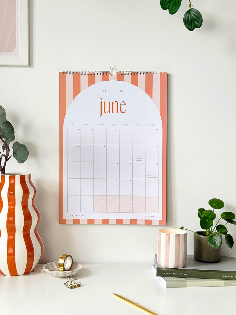 large A3 wall calendar with orange striped design showing june 2026 on a wall above a desk with decorative stationery items. 