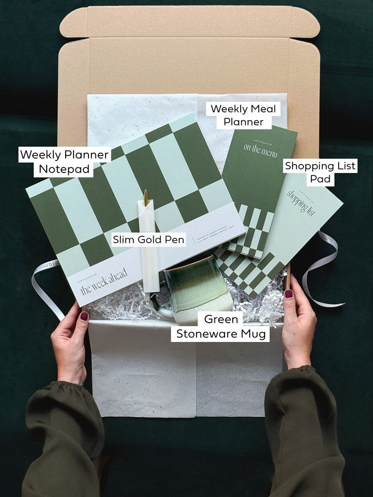 Labelled stationery items inside the Home Planning Set gift bundle: A4 weekly planner, meal planner, shopping list, gold pen and green stoneware mug.