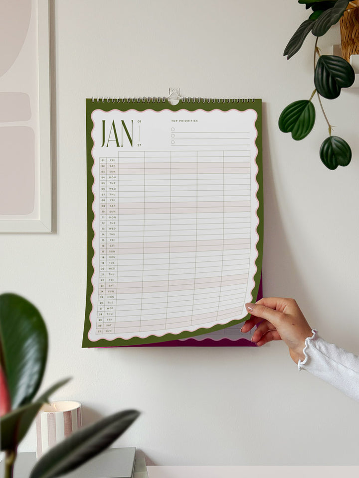 A January 2027 family wall calendar with a green scalloped border is hanging on a white wall, showing a vertical planner grid and 5 columns for each family member to add their plans.