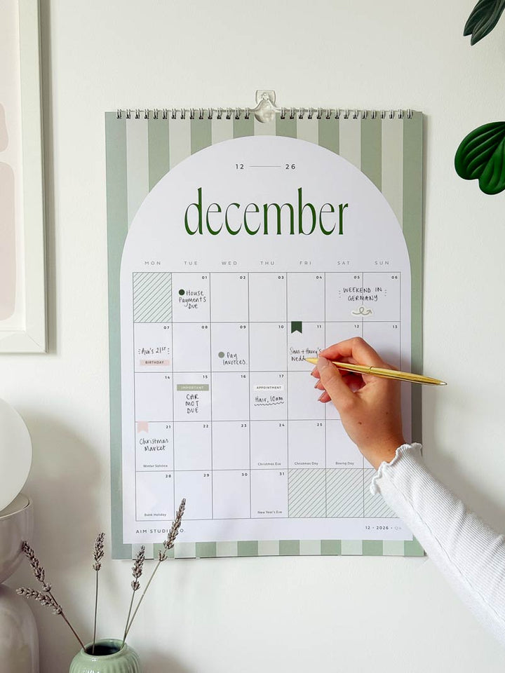 adding plans and appointments to a 2026 calendar hanging on a wall. the month view planner has handwritten appointments and cute stickers 