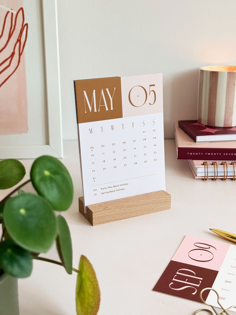 a small a6 desktop calendar displaying may 2027 month to view card in a wooden stand within a styled office desk scene.