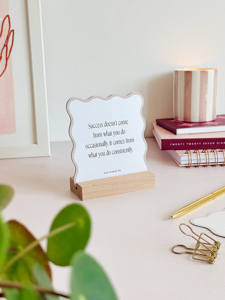 a positive affirmation card is held in a wooden stand on a light pink surface. the daily quote card mentions personal growth, success and consistency.