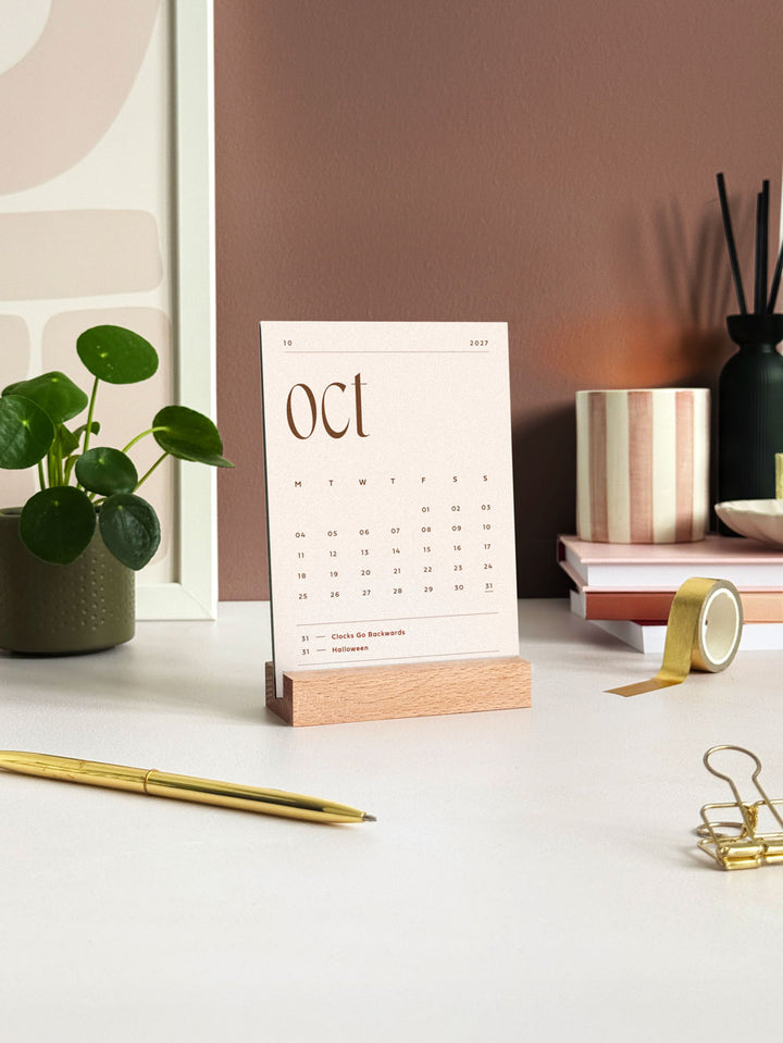 A monthly desk calendar showing October 2027 with a6-sized cards held in a wooden block stand. Styled on a minimal desktop with stationery ans gold accessories.