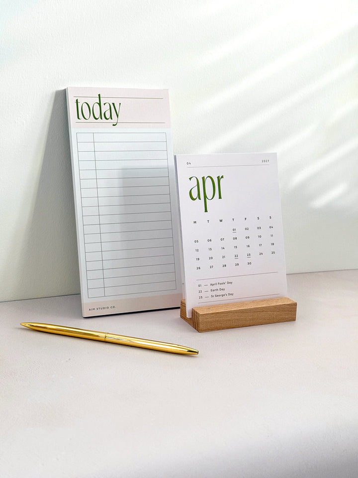 Pale pink daily time blocking notepad with a minimalist 2027 desk calendar in a wooden holder on a light office desk.
