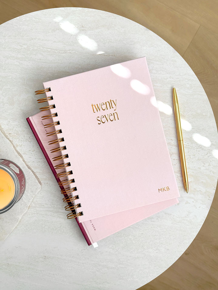 personalised 2027 spiral diary with pale pink cover and gold foiled intiails. Styled with a gold pen and candle on a white marble table.