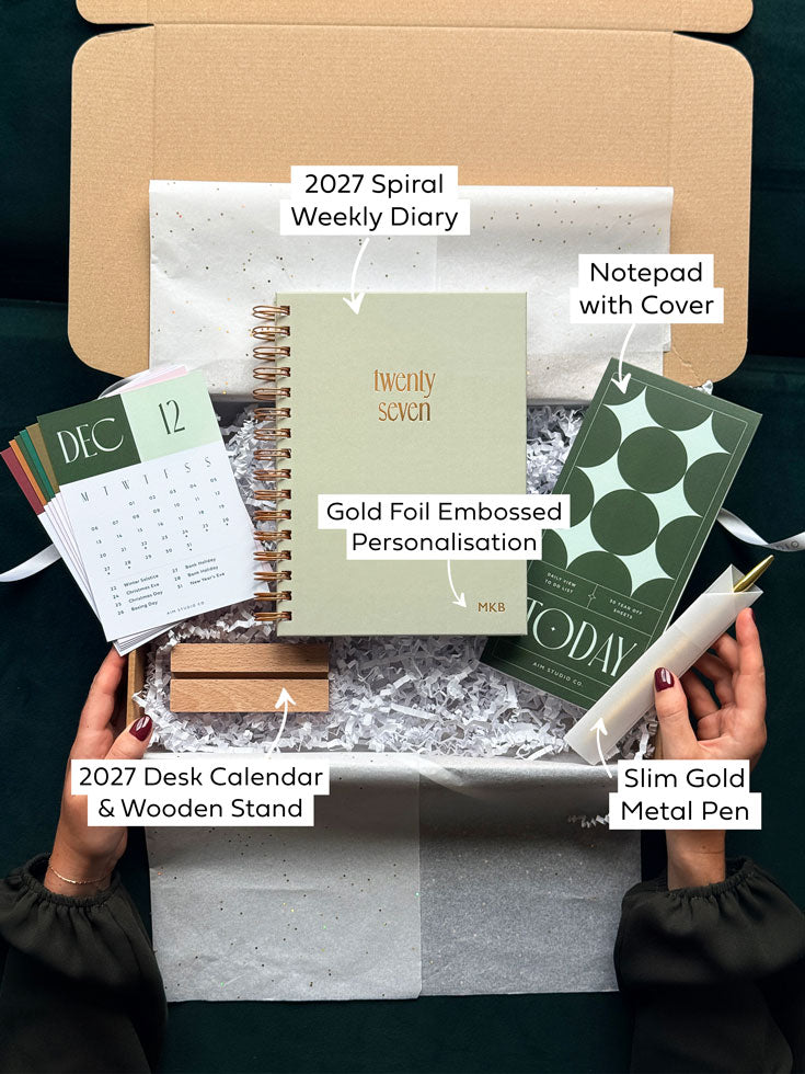 A curated gift box set opened by hands, featuring a 2027 spiral weekly diary, a notepad with a cover, a 2027 desk calendar with a wooden stand, a slim gold metal pen, and a gold foil embossed planner insert, arranged in white packing paper inside a brown box.