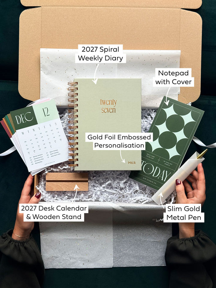 A curated gift box set opened by hands, featuring a 2027 spiral weekly diary, a notepad with a cover, a 2027 desk calendar with a wooden stand, a slim gold metal pen, and a gold foil embossed planner insert, arranged in white packing paper inside a brown box.