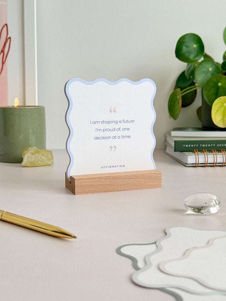 a wavy-edged affirmation card stands upright on an office desk, held in a wooden stand. Styled with other wavy cards in the foreground and candles and crystals in the background.