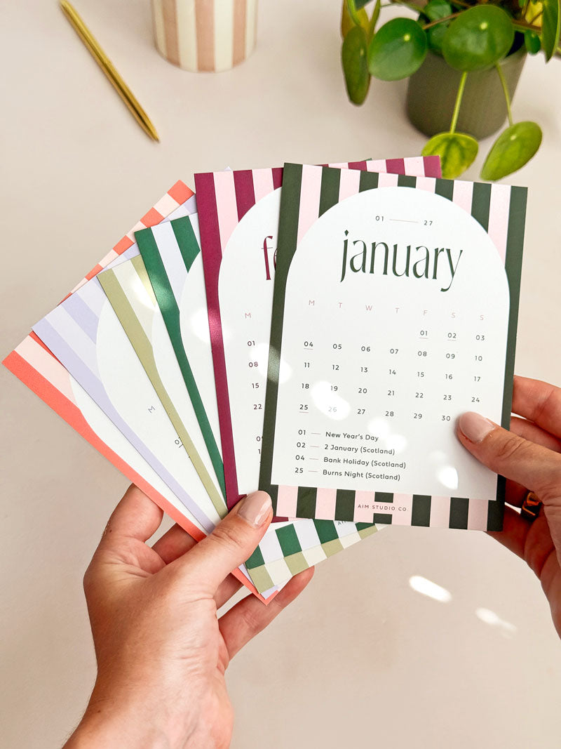 holding a set of striped monthly desk calendar cards with white arch containing date grid with UK bank holidays.