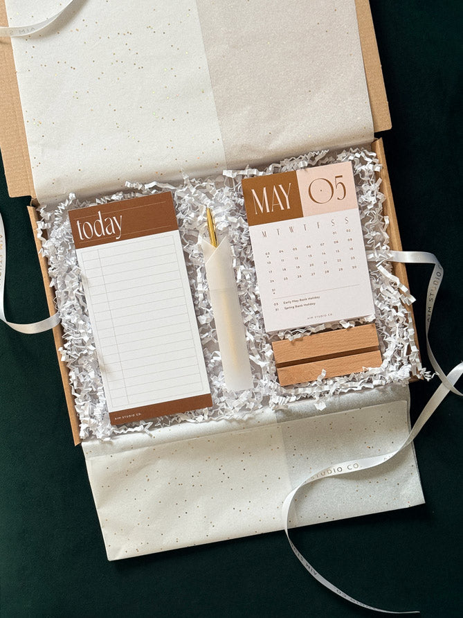 A kraft gift box with tissue paper lining and white crinkle paper filler, containing a lined notepad labeled &
