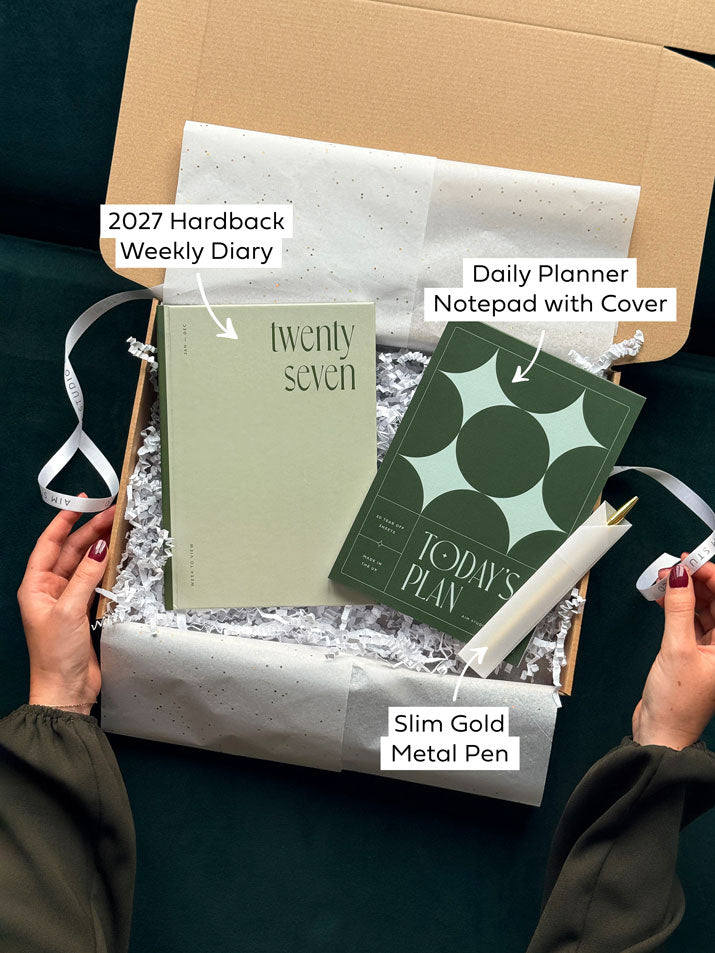 An open stationery gift set with two hands unwrapping. Inside the Christmas gift set there is a 2027 hardback weekly diary, daily planner notepad with dark green star cover and a slim gold metal pen. Text with arrows highlights each item of stationery included in the gift bundle.
