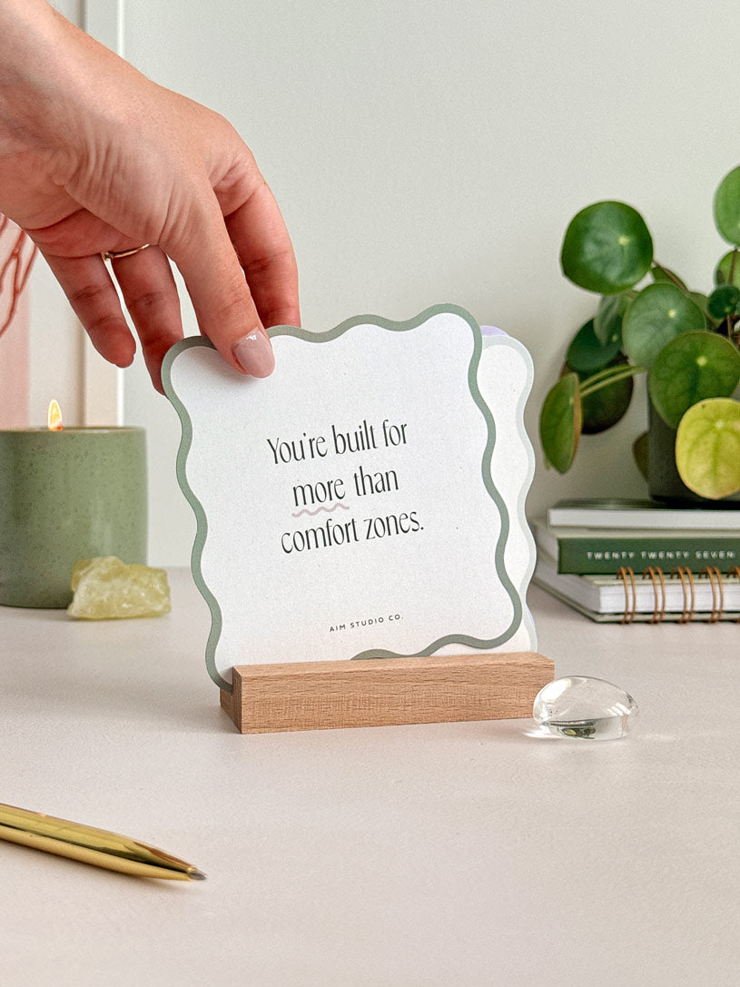 a woman is placing a mindfulness quote card which has a sage green wavy edges and modern serif typography. It is being placed into a wooden holder to display the motivational quote cards on an office desk.