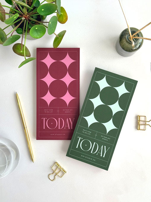 Two DL sized slim to do list planner pads on an office desk. Styled with aesthetic accessories like gold pen and paper clips, the notepads show the retro-inspired star design in two colour options: deep red and pink and deep green and pale green.