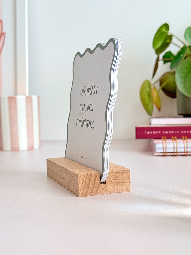 mindfulness card pack standing in a wooden holder on an office desk. The affirmation cards are cut into a wavy shape with a border highlighting the edges in a range of soft pastel colours. In the background there is a striped candle and a potted desk plant.