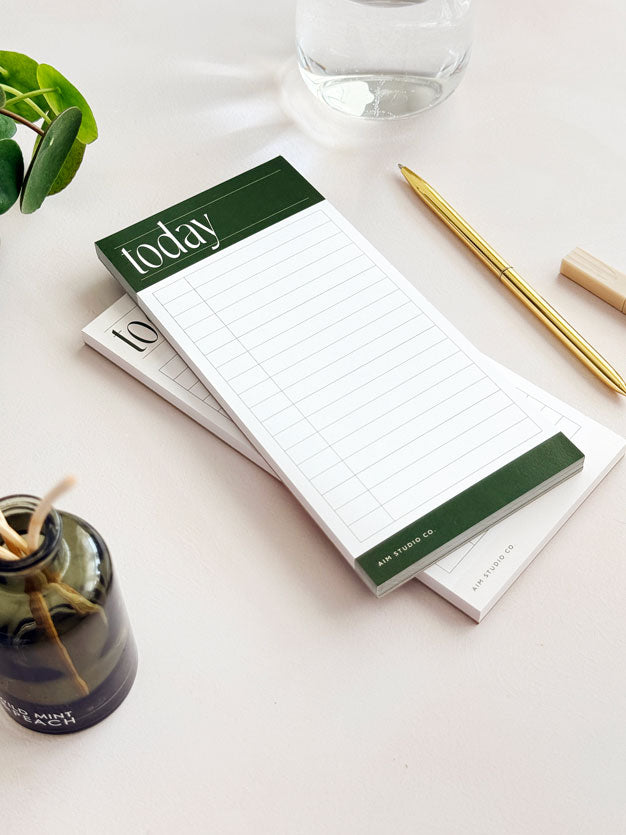 Notepad with green border labeled 'today' on a white surface with a plant and glass in the background.