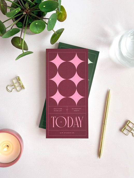 slim to do list notepad on an office desk with deep red and pink star cover. Styled with a gold pen and paper clips.
