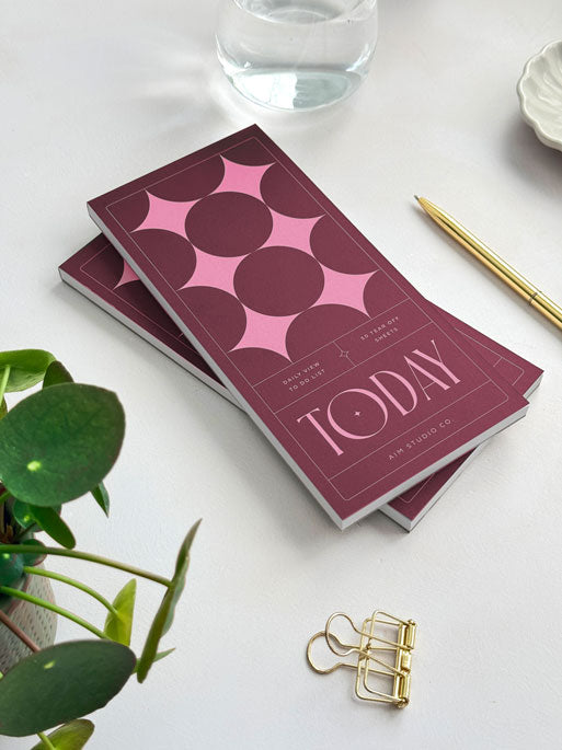 side view of DL-sized list pad with large serif typography on the cover reading "today" in pink. The cover has a matte finish and a deep red background with retro design and large pink stars. Surronded by desk accessories on a light surface.