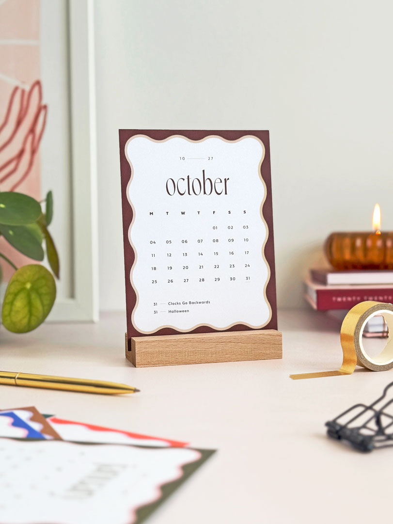 monthly desk calendar showing october 2027 with brown scalloped edge held in a natutal wooden stand. The small calendar is styled on an office desk with aesthetic stationery and desk accessories surrounding it.