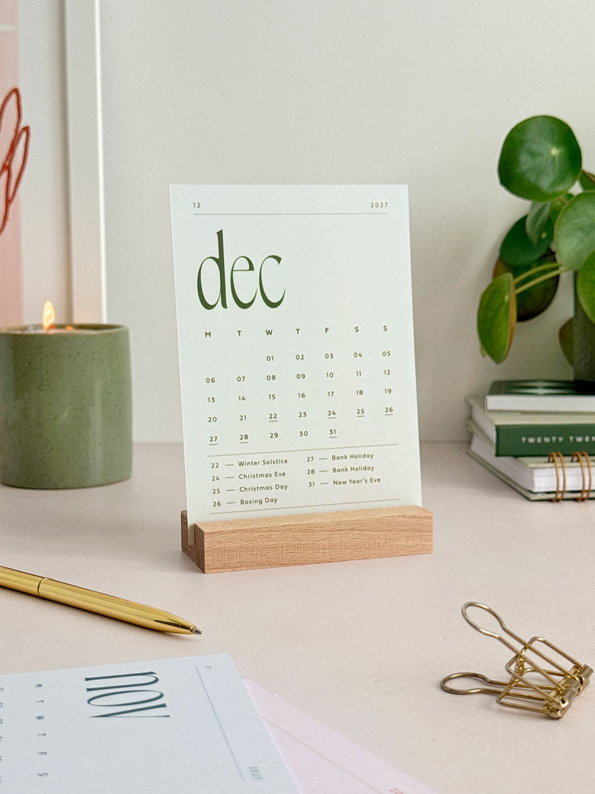 a monthly desk calendar displaying the calendar card of december 2027 with a sage green background and deep green text. The A6 card is displayed in a natural wooden stand on an office desk with aesthetic desk accessories.