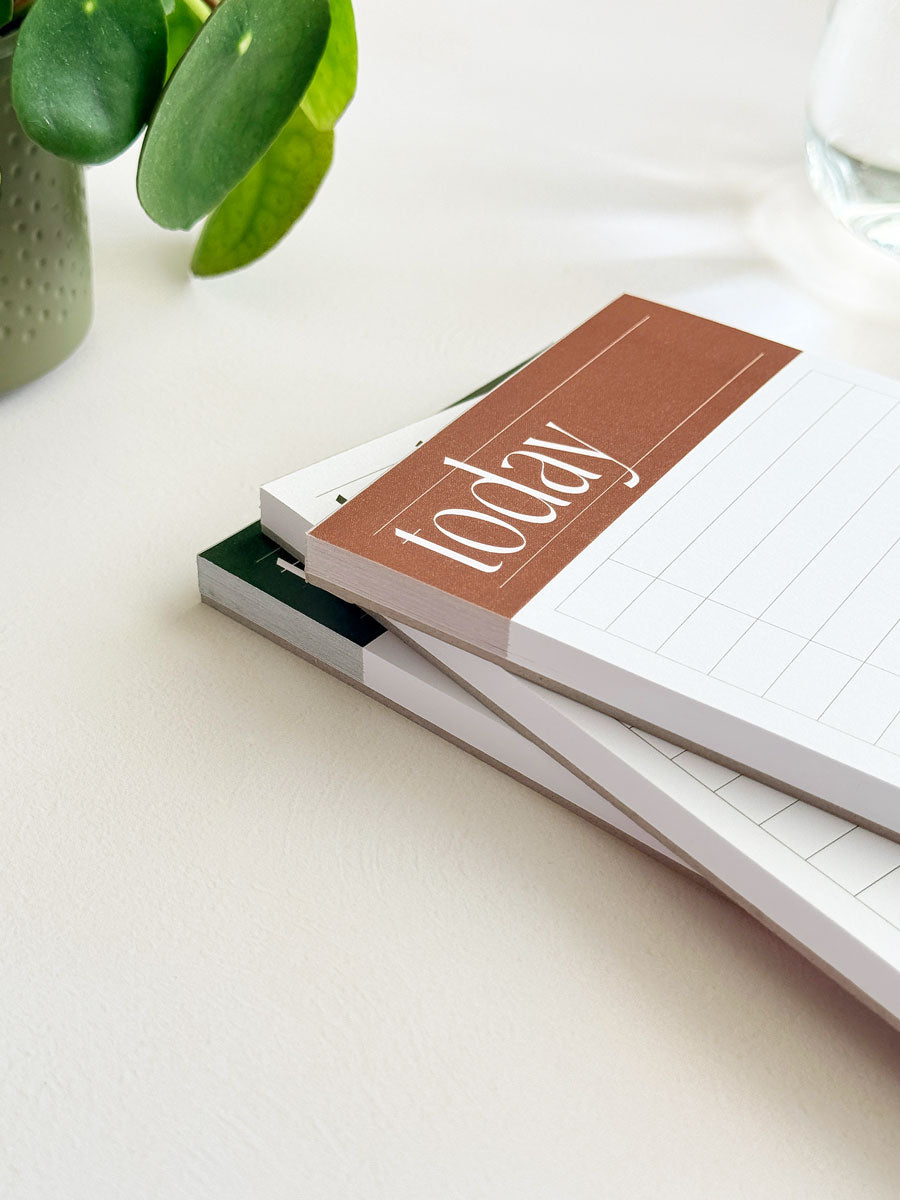 Stack of planner notepads with a 'today' cover on a light surface, with a plant in the background.
