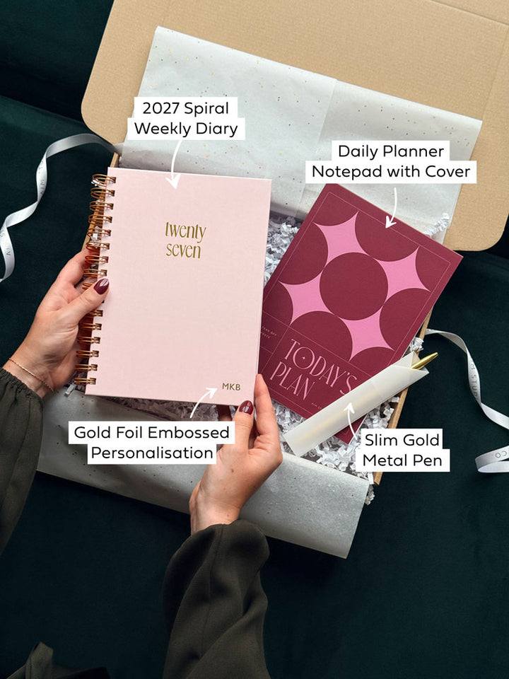 a gift set for her containing a 2027 spiral weekly diary, gold foiled cover with personalisation, a daily planner notepad with bold burgundy and pink cover and a slim gold pen. The stationery set is gift wrapped in white tissue paper, zig-zag shredded paper and tied with a branded ribbon.