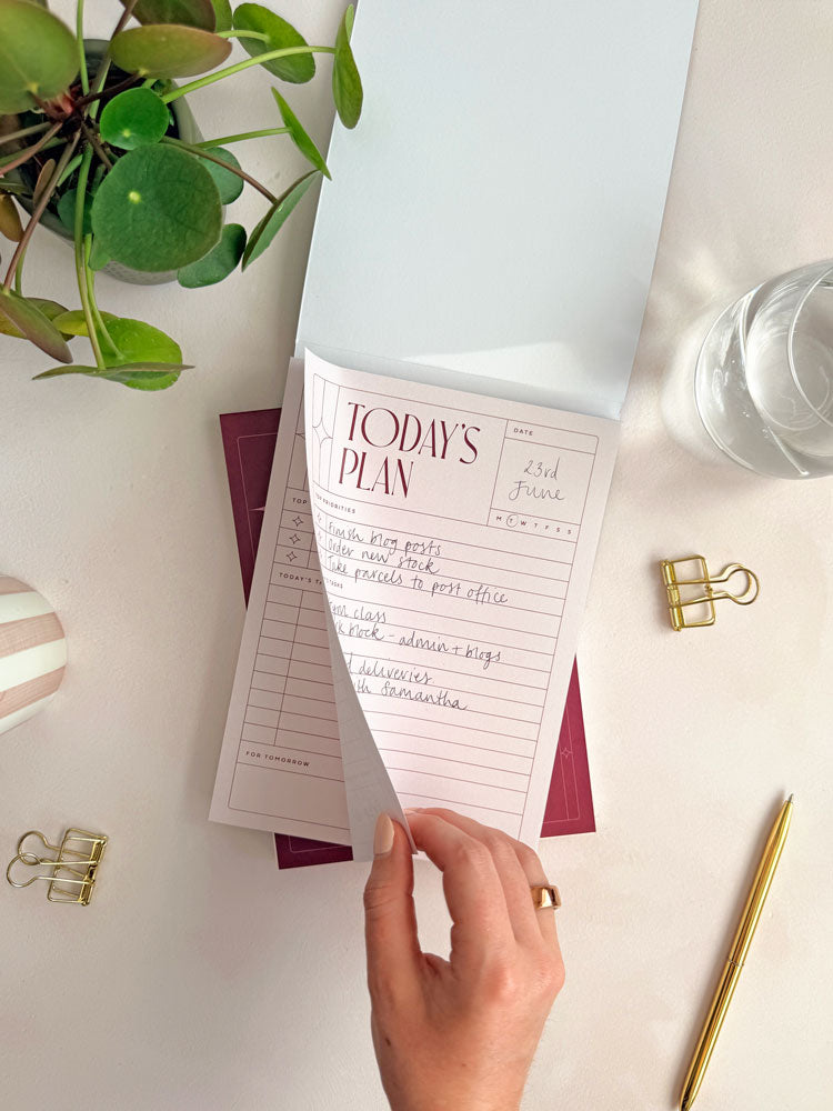 hand tearing off a daily planner page from a desk planner notepad with cover. Styled on a desktop surface with a glass of water, desk plant and office stationery.