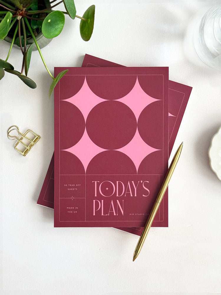 A styled flat lay of an A5 daily planner notepad with a deep burgundy cover and geometric stars in pink on the cover, titled "today&