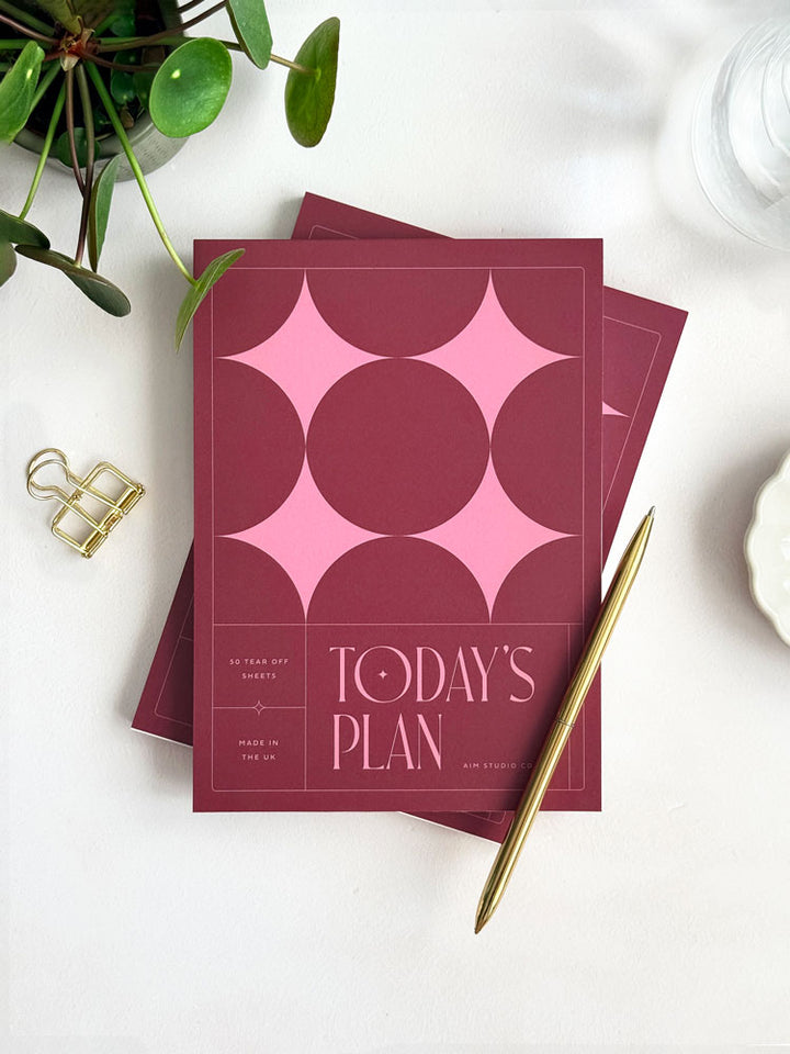 A styled flat lay of an A5 daily planner notepad with a deep burgundy cover and geometric stars in pink on the cover, titled "today&