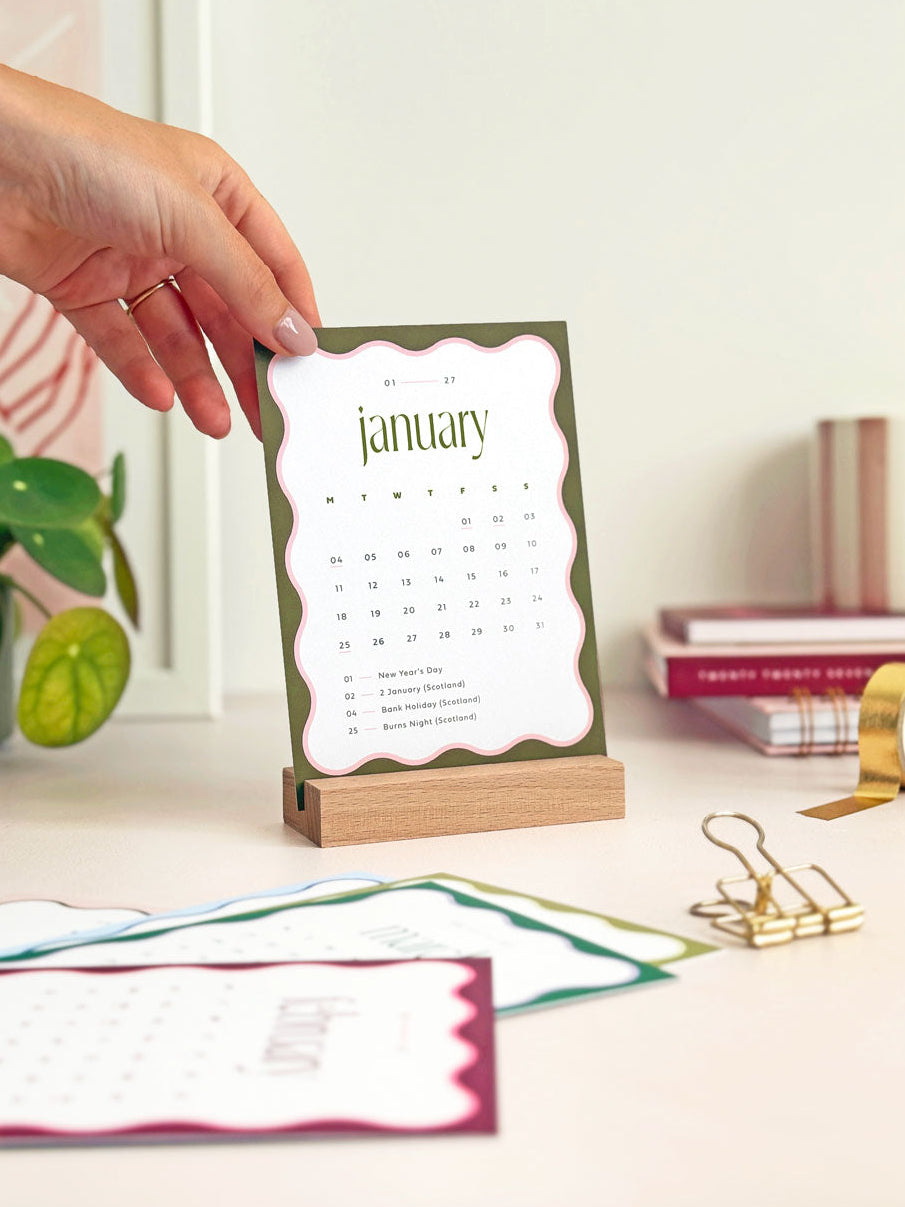 small desk calendar card displaying the month of january 2027 with deep green and pink wavy border design and minimal calendar grid with UK bank holidays.