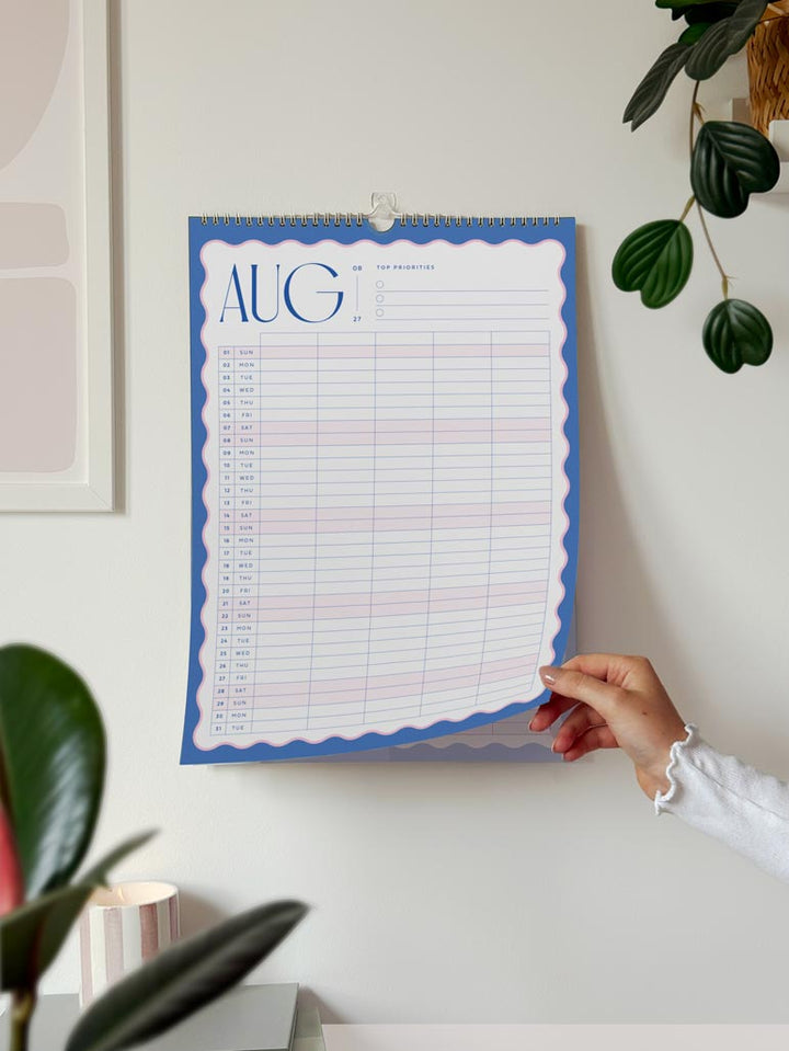 woman turning the page of large a3 wall planner for families showing the month of august 2027 with bright blue scalloped border and bold typography.