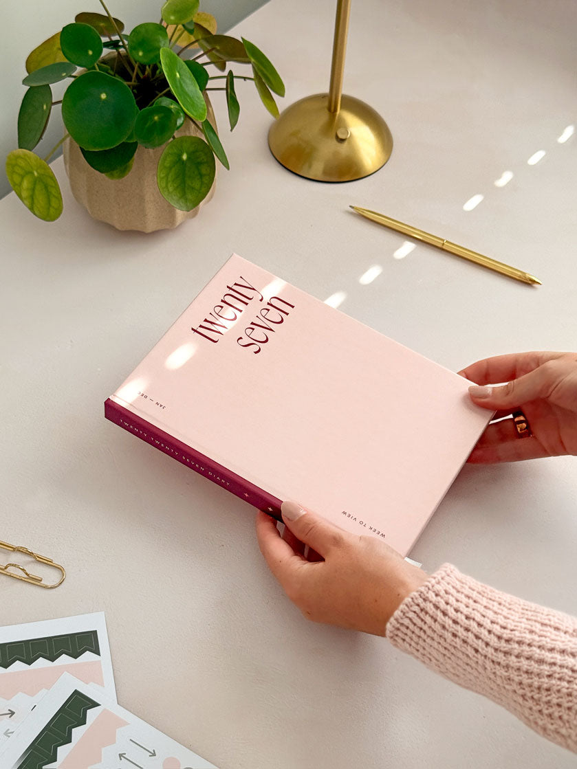 Woman placing a 2027 pink and red hardback planner on an office desk surrounded by planner sticker sheets, a gold pen, lamp and potted desk plant.